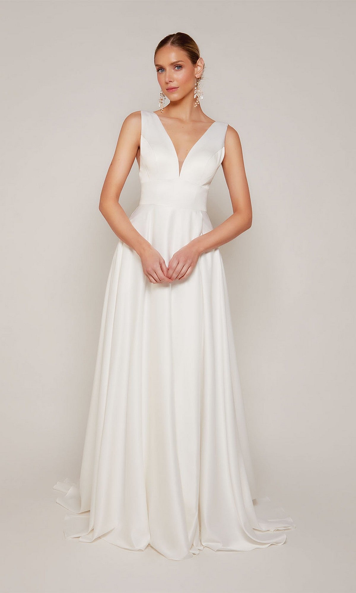 A woman wears the Long Formal Dress: Alyce 7096, a sleeveless, floor-length white gown featuring a deep V-neckline and elegant A-line silhouette, standing against a plain light background.